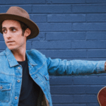 A man with a hat and a guitar leaning against a brick wall.