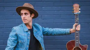 A man with a hat and a guitar leaning against a brick wall.