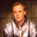 A man in a silver jacket sitting in a dark room.