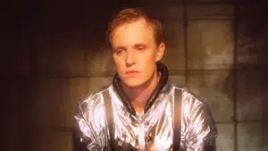 A man in a silver jacket sitting in a dark room.