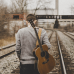 A man standing on railroad tracks with an acoustic guitar.