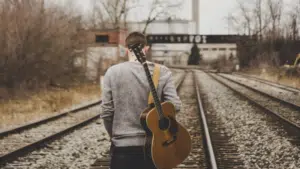 A man standing on railroad tracks with an acoustic guitar.