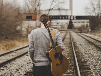 A man standing on railroad tracks with an acoustic guitar.