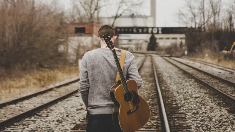 A man standing on railroad tracks with an acoustic guitar.