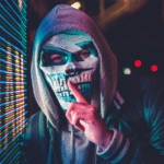 A man in a hoodie with a mask posing as an anonymous artist in front of a screen.