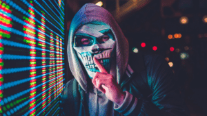 A man in a hoodie with a mask posing as an anonymous artist in front of a screen.