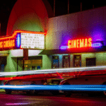 Neon-lit cinema with a marquee displaying film titles, ideal for a sync licensing backdrop. Parked cars line the street as light trails from passing traffic paint the foreground.