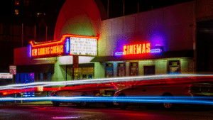 Neon-lit cinema with a marquee displaying film titles, ideal for a sync licensing backdrop. Parked cars line the street as light trails from passing traffic paint the foreground.