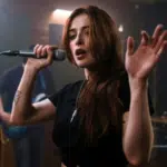 A woman with long hair sings into a microphone, raising her hand in an electrifying moment captured perfectly for press photos. Musicians with instruments play energetically in the background.