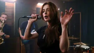 A woman with long hair sings into a microphone, raising her hand in an electrifying moment captured perfectly for press photos. Musicians with instruments play energetically in the background.