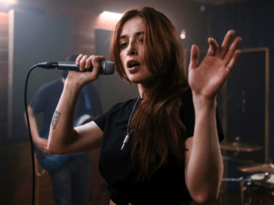 A woman with long hair sings into a microphone, raising her hand in an electrifying moment captured perfectly for press photos. Musicians with instruments play energetically in the background.