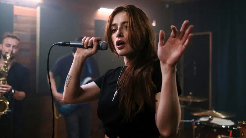 A woman with long hair sings into a microphone, raising her hand in an electrifying moment captured perfectly for press photos. Musicians with instruments play energetically in the background.