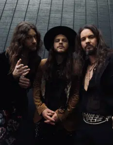 Three individuals with long hair and beards, dressed in eclectic, stylish clothing, pose against a dark textured wall for their press photos.