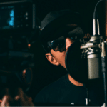 A person wearing sunglasses and a hat is speaking into a microphone in a dimly lit room with professional recording equipment, reminiscent of an artist working under indie labels.