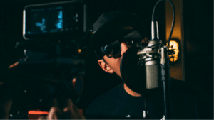 A person wearing sunglasses and a hat is speaking into a microphone in a dimly lit room with professional recording equipment, reminiscent of an artist working under indie labels.