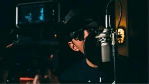 A person wearing sunglasses and a hat is speaking into a microphone in a dimly lit room with professional recording equipment, reminiscent of an artist working under indie labels.