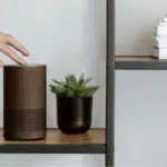 A person is pointing to an amazon echo speaker on a shelf, showcasing its voice capabilities.