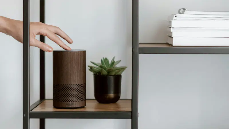 A person is pointing to an amazon echo speaker on a shelf, showcasing its voice capabilities.