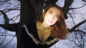 A person with red hair and light skin wearing a black top with lace trim stands outdoors in front of a large tree.