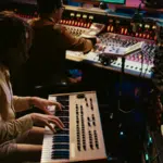 In a cozy music studio, two artists create magic: one immerses in the keyboard synthesizer while the other perfects mixing console settings. Equipment and screens surround them as they craft tracks ready to captivate listeners on Bandcamp.