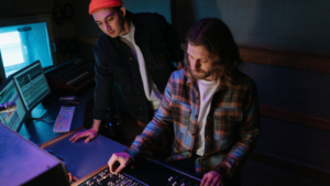 Two men work in a dimly lit recording studio, one focused on management as he adjusts controls on a mixing console while the other observes.