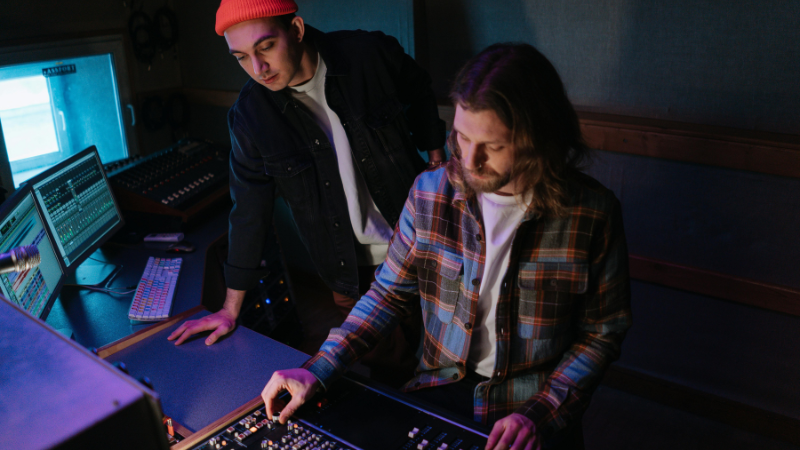 Two men work in a dimly lit recording studio, one focused on management as he adjusts controls on a mixing console while the other observes.