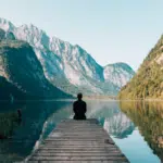 A man peacefully meditating on a dock in front of a serene mountain lake.