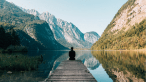 A man peacefully meditating on a dock in front of a serene mountain lake.