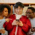 Three people stand in front of a bookshelf, looking at a phone held by the person in the center. The people on the sides are holding phones and appear engaged as they plan their social media content calendar.