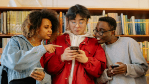 Three people stand in front of a bookshelf, looking at a phone held by the person in the center. The people on the sides are holding phones and appear engaged as they plan their social media content calendar.