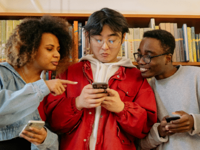 Three people stand in front of a bookshelf, looking at a phone held by the person in the center. The people on the sides are holding phones and appear engaged as they plan their social media content calendar.