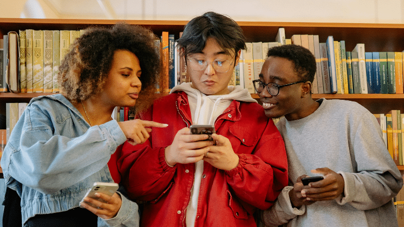 Three people stand in front of a bookshelf, looking at a phone held by the person in the center. The people on the sides are holding phones and appear engaged as they plan their social media content calendar.