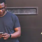 A man is browsing Clubhouse on his phone in front of a brick wall.