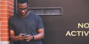 A man is browsing Clubhouse on his phone in front of a brick wall.