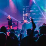 A crowd enjoys a live music performance with vibrant stage lighting and a DJ in the background, while raised hands indicate enthusiastic fan engagement.