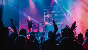 A crowd enjoys a live music performance with vibrant stage lighting and a DJ in the background, while raised hands indicate enthusiastic fan engagement.