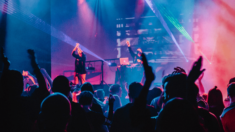 A crowd enjoys a live music performance with vibrant stage lighting and a DJ in the background, while raised hands indicate enthusiastic fan engagement.