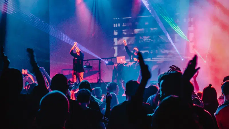 A crowd enjoys a live music performance with vibrant stage lighting and a DJ in the background, while raised hands indicate enthusiastic fan engagement.