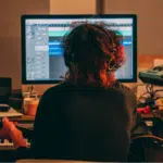 Person with headphones sits at a desk with a computer screen displaying audio editing software, holding a guitar, surrounded by musical equipment and keyboards, preparing to streamline his next project that could significantly boost his revenue.