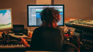 Person with headphones sits at a desk with a computer screen displaying audio editing software, holding a guitar, surrounded by musical equipment and keyboards, preparing to streamline his next project that could significantly boost his revenue.