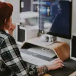 A person with red hair in a plaid shirt is sitting at a desk, using a computer. The desk has a raised monitor, keyboard, speakers, clock, and other office equipment, creating the perfect setup for managing copyright tasks efficiently.