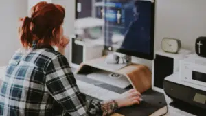 A person with red hair in a plaid shirt is sitting at a desk, using a computer. The desk has a raised monitor, keyboard, speakers, clock, and other office equipment, creating the perfect setup for managing copyright tasks efficiently.