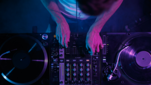 A person operates a DJ mixer and two turntables under purple and blue lighting, adjusting controls and playing vinyl records—showcasing essential music promotion tips in action.