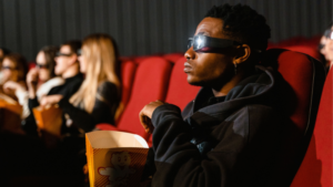 A group of people wearing 3D glasses sit in red theater seats, watching a movie in sync and eating popcorn.