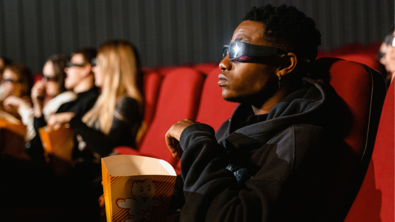 A group of people wearing 3D glasses sit in red theater seats, watching a movie in sync and eating popcorn.