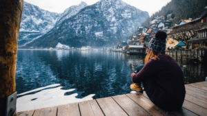 As the music industry slowdown reaches its peak, a woman finds solace sitting on a dock overlooking a serene lake with majestic mountains in the background.
