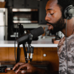 A man in headphones is sitting in front of a microphone, surrounded by his back catalog.