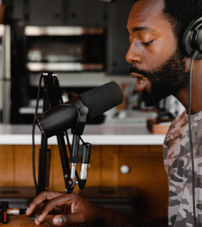 A man in headphones is sitting in front of a microphone, surrounded by his back catalog.