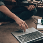 A person is comfortably sitting on a sofa, laptop balanced nearby and a guitar beside them, with headphones resting around their neck, ready to dive into the world of SplitShare for seamless creative sharing.