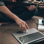 A person is comfortably sitting on a sofa, laptop balanced nearby and a guitar beside them, with headphones resting around their neck, ready to dive into the world of SplitShare for seamless creative sharing.